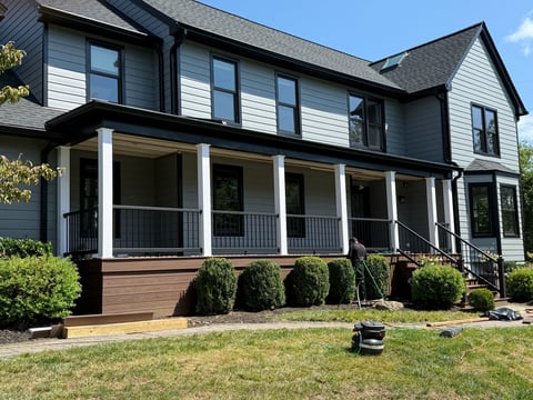 Modern two-story house with gray and white exterior, wraparound porch with white columns, dark roof, and manicured landscaping in front yard