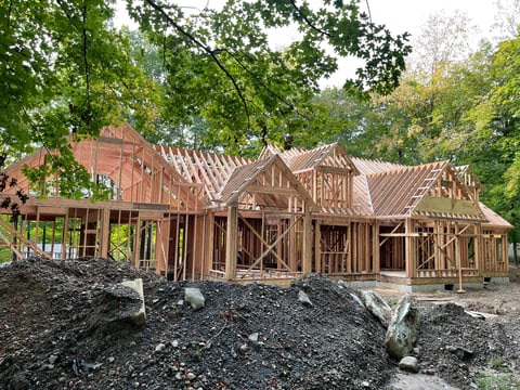Large house under construction with wooden frame and brick exterior, surrounded by green trees and construction materials