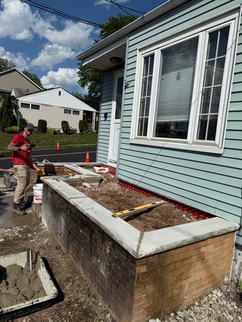 Worker in red shirt building a brick and concrete foundation against a light blue house with large windows