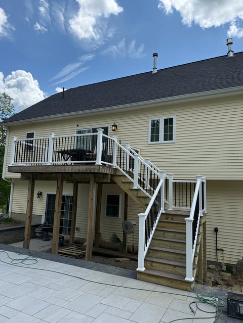 Two-story house with cream-colored siding, dark roof, and white wooden deck with stairs against blue sky