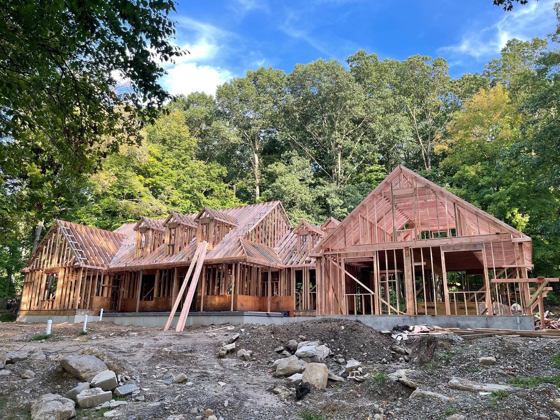 Large wooden house under construction with exposed framing, surrounded by green trees and rocky ground